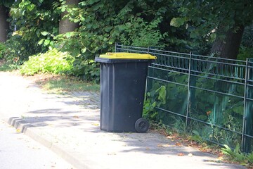 dark plastic garbage can with yellow lid is ready for collection at the roadside at the fence