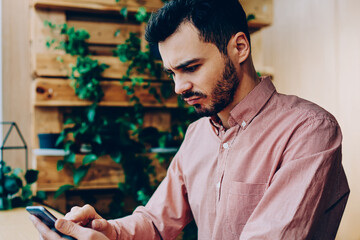 Serious young male freelancer disappointed with bad news while checking mail on smartphone sitting in coworking space,stressed hipster guy angry about reading banking balance via app on cellphone