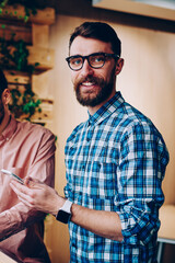 Cheerful male it developer looking at camera and holding telephone while his friend reading information from web on laptop computer, prosperous employee cooperating with colleague on project