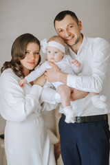 Obraz premium A family of three, a man and two women, are holding a baby. The baby is wearing a white dress and a white headband. The family is smiling and he is happy