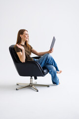Happy woman sitting in armchair with tablet having online meeting against white studio background.