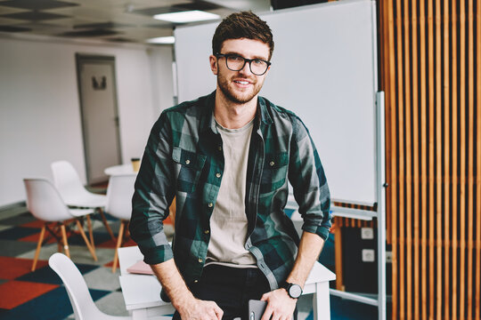 Half length portrait of handsome successful male designer wearing glasses for provide eyes protection enjoying break time at work and looking at camera while sitting on table in modern office