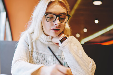 Clever female student in spectacles writing homework task in notepad sitting with laptop computer...
