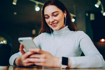 Cheerful teen girl watching favorite serial on smartphone via good wifi connection in cafe, smiling young woman using cellular for downloading new songs for playlist listening music on coffee break