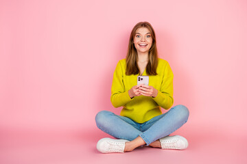 Young Smiling Woman Holding Smartphone Against Pink Background in Casual Yellow Sweater and Jeans