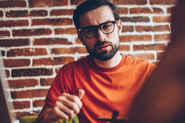 Sceptical bearded young man in optical eyeglasses looking at modern laptop computer device connected to 4G internet and thinking on technical problem sitting in coworking space interior