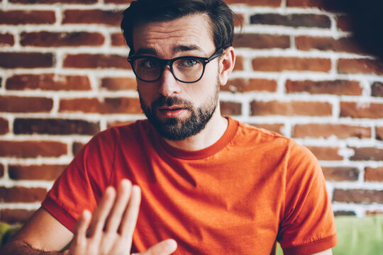Sceptical hipster guy dressed in trendy orange t-shirt arguing during meeting.Young man with beard and eyeglasses for vision correction showing sign stop sitting in stylish coworking space