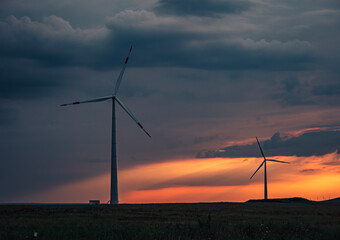 Obraz premium Wind turbines silhouetted against a vibrant sunset over rolling fields in rural landscape