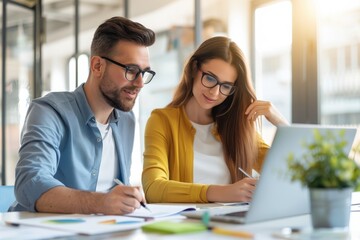 Obraz premium Two young professionals collaborate on a laptop in a bright office, taking notes and brainstorming ideas, showcasing teamwork and productivity.