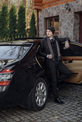 A man in a suit and hat stands next to a black car. He is holding a drink and he is waiting for...