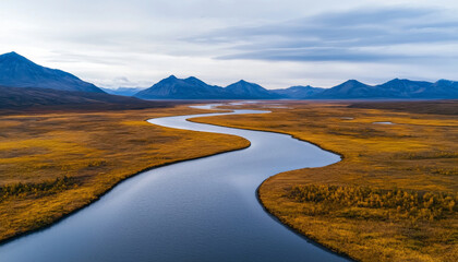 Fototapeta premium Winding river through golden grasslands and mountains under cloudy sky