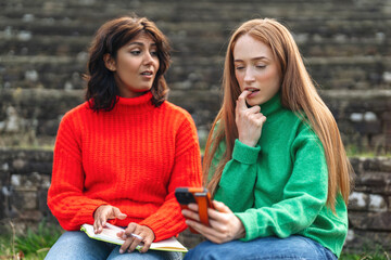 Two friends engaged in conversation while sitting on outdoor steps in colorful sweaters during a sunny day