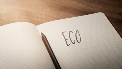 A handwritten inscription "ECO" on a grille of an open notebook on a wooden countertop, next to a black pencil, lighting of light. (selective focus)