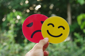 Hand holding a sad face card hiding behind a happy smiling face. Bipolar and depression concept, mental health concept, personality, mood swings