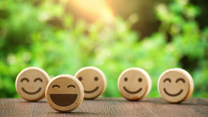 Five smiling faces carved on a round wooden surface with a background of blurred lush green leaves. A cheerful and optimistic concept, conveying a sense of joy and positive emotions.