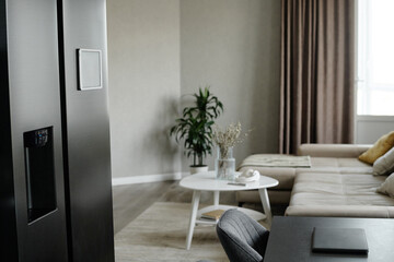 Smart fridge next to dining table in kitchen zone, spacious and minimalist style living room with beige sofa and wooden coffee table