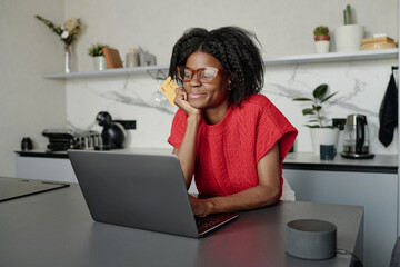 Attractive African American housewife leaning on bar counter while shopping online and holding tightly her credit card