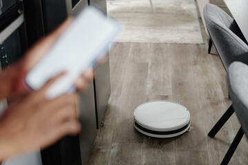 Blurred hands controlling white robot vacuum cleaner via smartphone application while robot vacuum cleaning parquet, focus on robot vacuum cleaner
