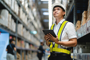 A man wearing a yellow vest and a hard hat is holding a tablet in a warehouse. He is looking up at something on the tablet