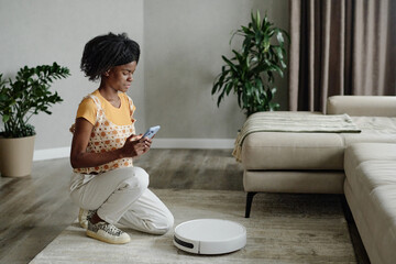 African American housewife squatting while turning on robot vacuum cleaner in living room