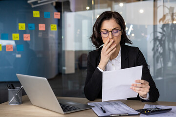 Businesswoman at office desk reviews financial documents with laptop. Shows emotion of stress,...
