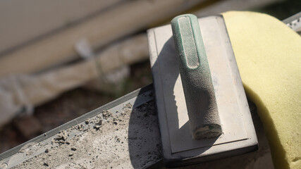 Closeup of a green plastic trowel and a yellow sponge resting on the edge of a cement wall after use. Concept of working equipment in a construction site