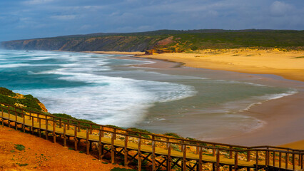 Dramatic Coastal Scenery in Algarve: Overcast Sky, Wooden Boardwalk Along Sandy Beach, Vibrant Waves, and Green Cliffs Capturing the Natural Beauty of Portugal's Shoreline