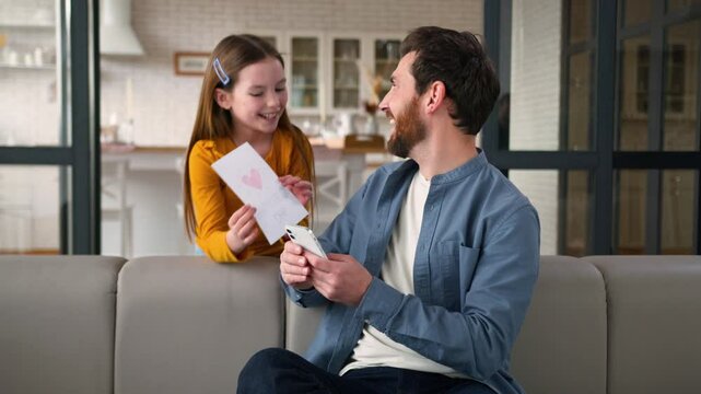 Carefree Caucasian girl kid surprise guy man father child schoolgirl giving handmade card present happy family rejoicing at home daughter congratulate hug parent dad holiday Father's Day male smiling