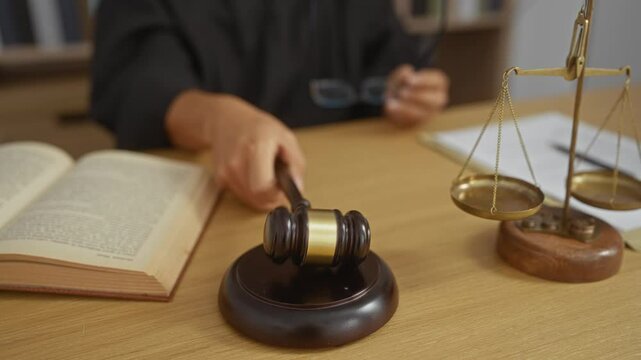 Judge in courtroom with gavel, law book, and scales of justice on wooden desk, indicating legal authority and decision making in formal judicial setting.