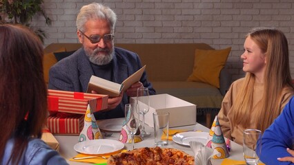 Family sit at table at home, grandchildren and parents celebrating birthday of grandfather, grandpa got a present box with new book.