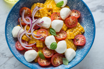 Salad with corn, red cherry tomatoes, red onion and mini mozzarella served in a blue bowl, horizontal shot, close-up
