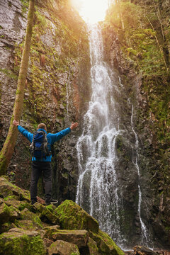 Tourist attraction of Germany - falls of Burgbach Waterfall near Schapbach, Black Forest, Baden-Wurttemberg, Germany. Man hiker in blue jacket standing on stone and looks at flow of falling water