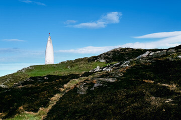 The image shows a white conical structure on a hill. The hill is a mix of grass and dark, rocky patches. The sky is mostly clear and blue.