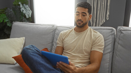 Fototapeta premium Young man indoors at home with beard relaxing on sofa using tablet in cozy living room with pillows and decorative plants enjoying a casual moment in a modern apartment setting.