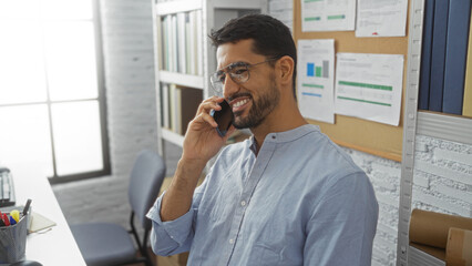 Young man with beard talking on phone in a bright office setting, surrounded by shelves and bulletin board, wearing glasses and smiling confidently.