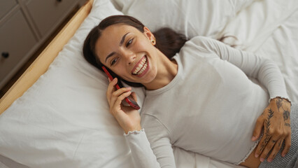 Young woman talking on a phone while lying on a bed with a cheerful expression in a cozy bedroom