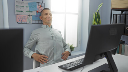 Fototapeta premium Young man relaxing in office setting, taking break from computer work, seated at desk in modern workplace, hands resting on waist, pleasant demeanor, bright interior, indoor environment