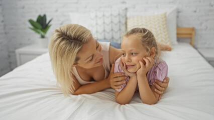 Blonde woman and her young daughter lying on a bed together at home in a cozy bedroom interior, sharing a tender moment.