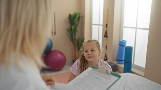 Woman therapist consulting child patient in modern indoor physio clinic, with therapy equipment visible in the room.