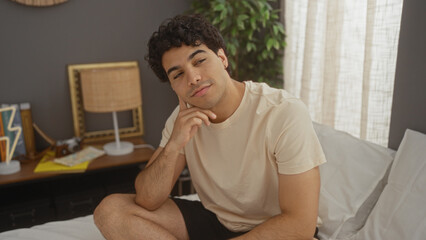 Fototapeta premium A handsome, young hispanic man sits thoughtfully on a bed in a well-decorated bedroom, featuring lamps and plants, creating a cozy and attractive indoor atmosphere.