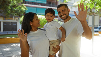 Latin family with woman, man, and child smiling and waving in an urban outdoor park setting during daylight, demonstrating love and togetherness.