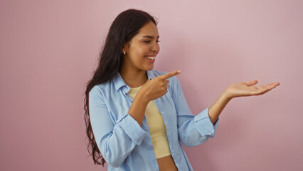 Beautiful young woman pointing at her extended hand over an isolated pink background, showing a product or idea with a happy smile.