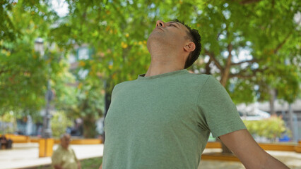 Handsome hispanic man relaxing outdoors in a green city park with trees in the background and a mature person sitting on a bench