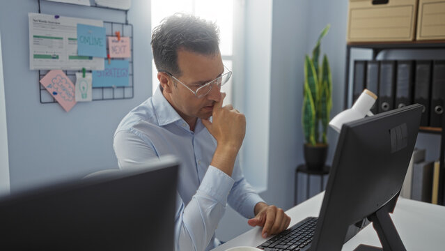 Hispanic man working in an office environment, thoughtfully looking at the computer screen with a cork board in the background and office plants nearby.
