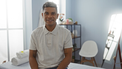 Young hispanic man sitting indoors at a rehab clinic workplace, smiling confidently in a relaxed interior setting with towels and a shelf in the background.