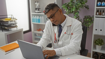 Obraz premium Hispanic male doctor working on a laptop in a clinic, managing phone call and looking focused in an indoor medical office setting.