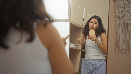 Young hispanic woman singing with hairbrush in hand while looking in the mirror in a wellness room...