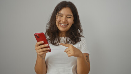 A young hispanic brunette woman pointing at a red smartphone, smiling, and looking attractive against an isolated white background wall.