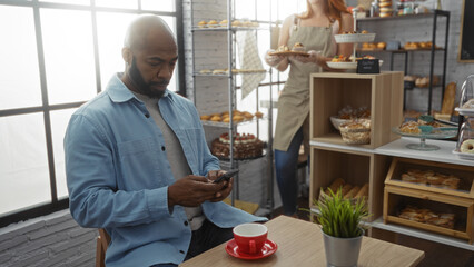 Man sitting in a bakery checking his phone while a woman baker serves pastries in the background, creating a cozy and inviting atmosphere indoors.