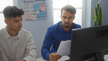 Two hispanic men working together in an office, focused on documents and computer screens, embodying the professional environment with teamwork and collaboration.